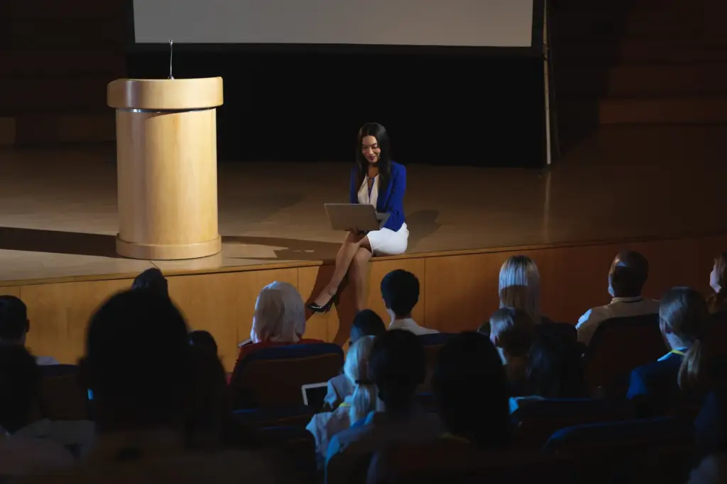 A woman in a blue jacket sits on the edge of a wooden stage with a laptop, beside a lectern, with the audience seated in front of her