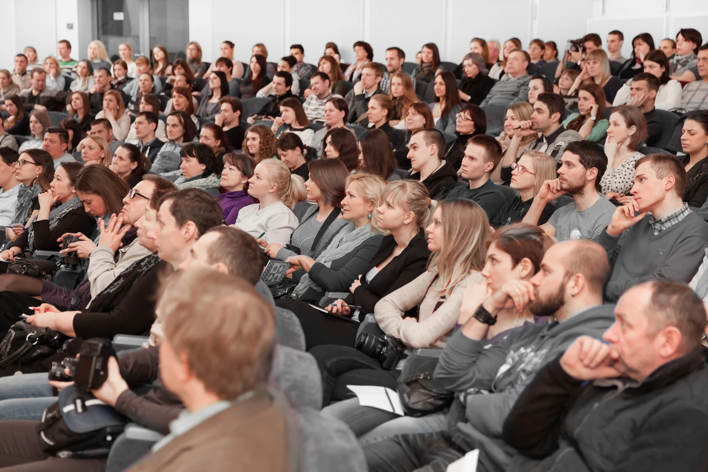 Rows of seated guests listening attentively in a bright auditorium, several taking notes or holding cameras