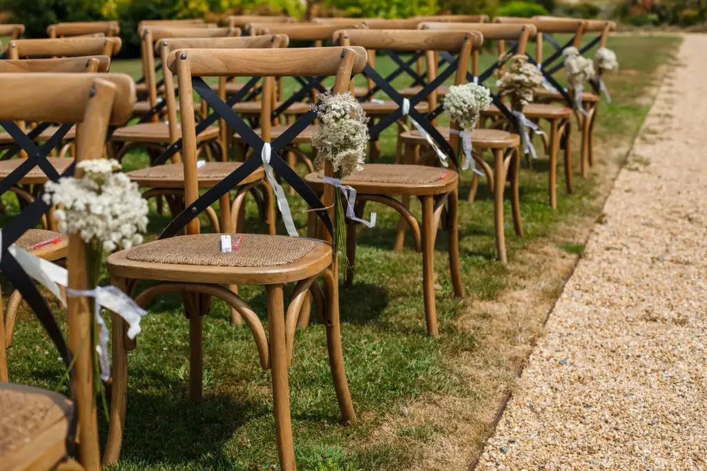 Rows of wooden chairs set out on grass beside a gravel path, with small white flower posies tied to the aisle seats