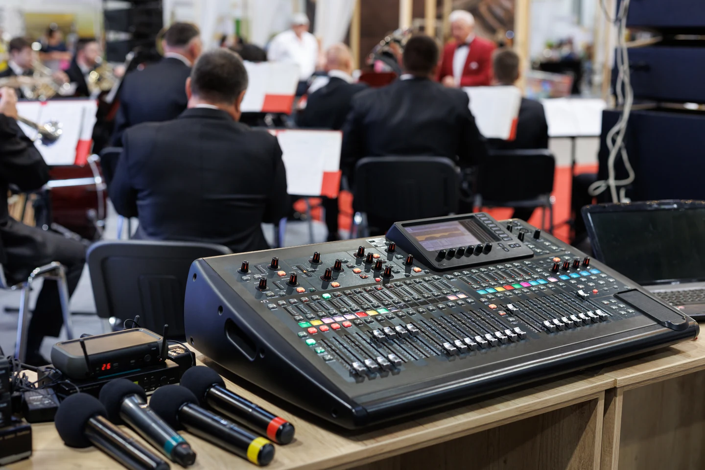 A digital audio mixing console and three wireless handheld microphones on a table, with a seated brass ensemble out of focus behind