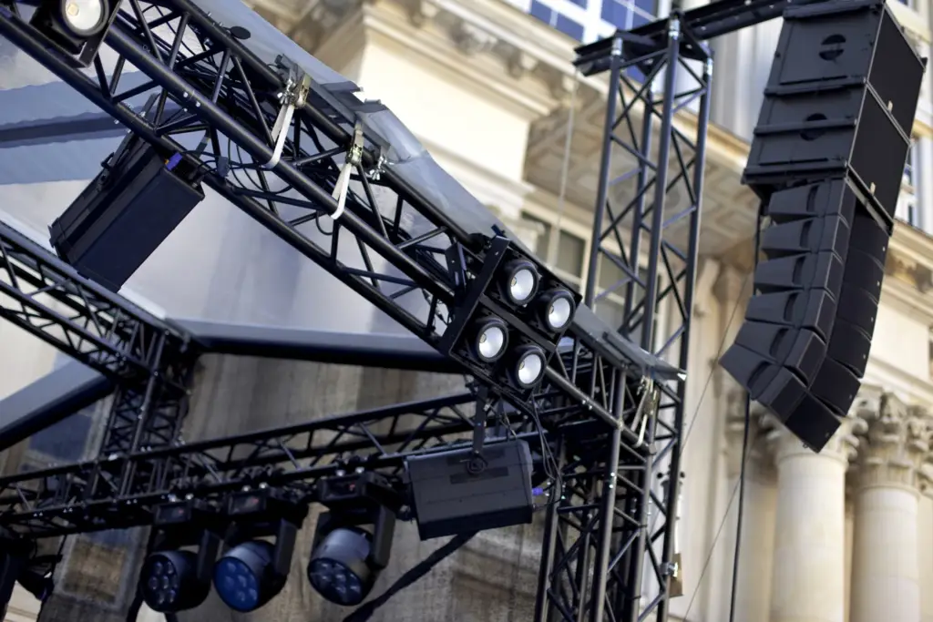 Stage lighting truss and line-array speakers rigged on an outdoor stage in front of a stone building