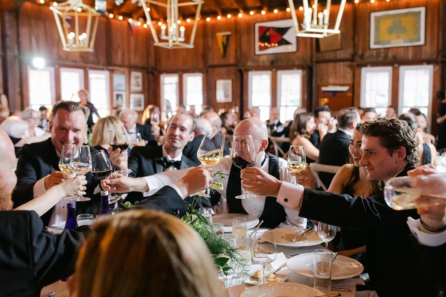 Wedding guests raising glasses in a toast at round tables in a timber hall lit by brass pendants and string lights