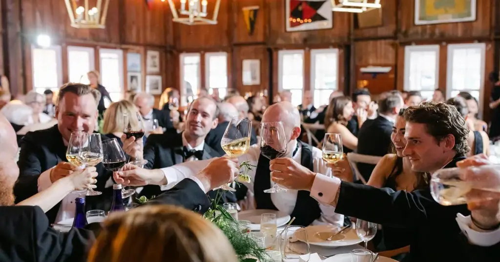 Wedding guests raising glasses in a toast at round tables in a timber hall lit by brass pendants and string lights